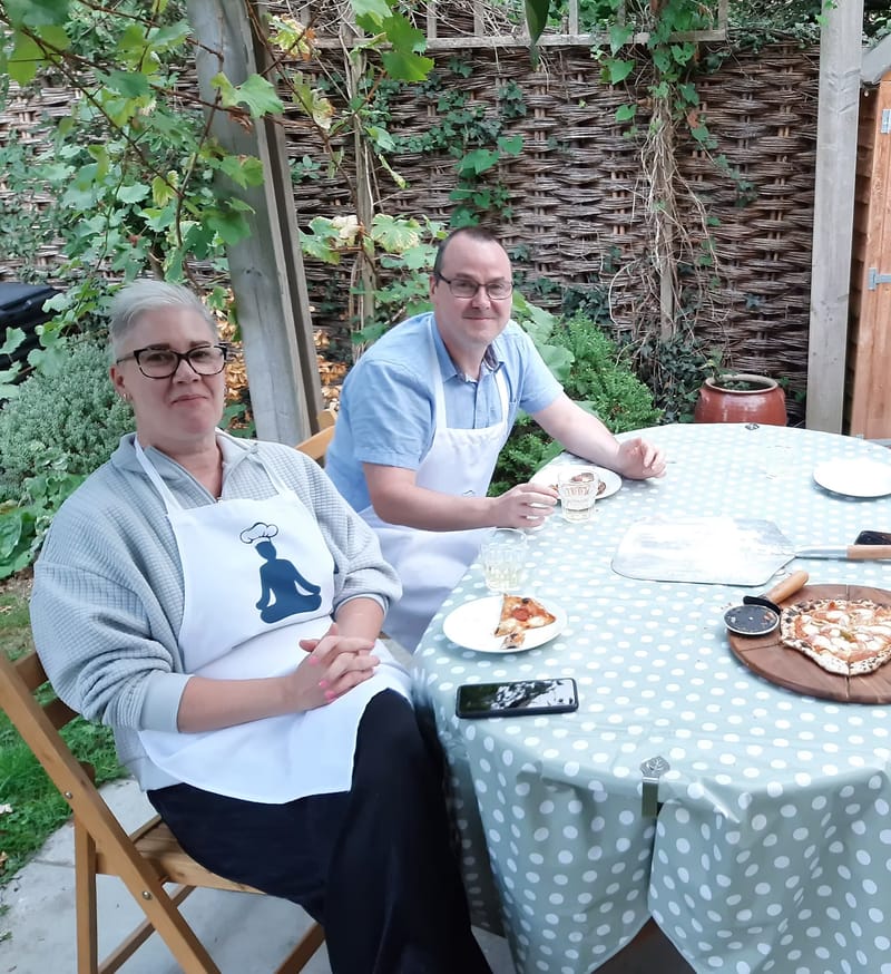 Two grown-up guests in aprons at a garden table with freshly baked pizza and a pizza peel