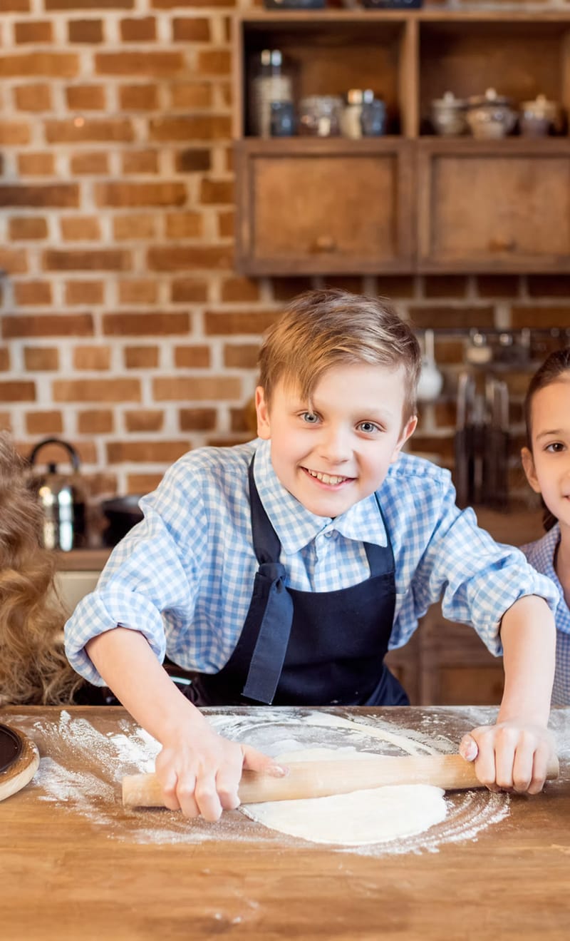 A smiling boy in a navy apron rolling out pizza dough on a floury wooden worktop