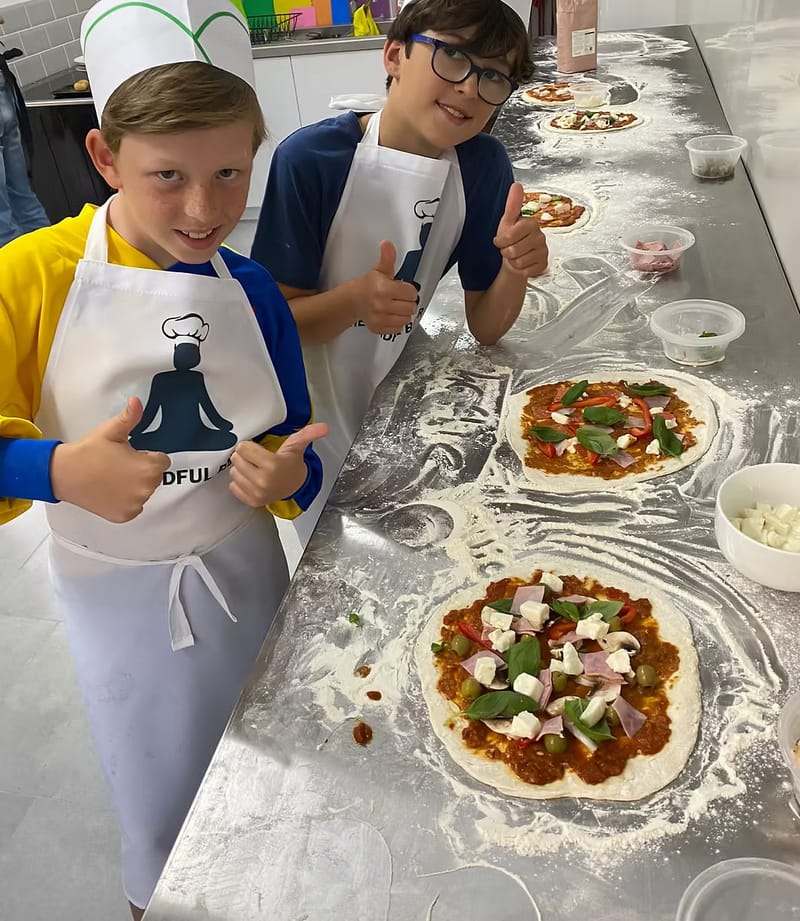 Two boys in Mindful Baker aprons giving a thumbs up beside the pizzas they've topped