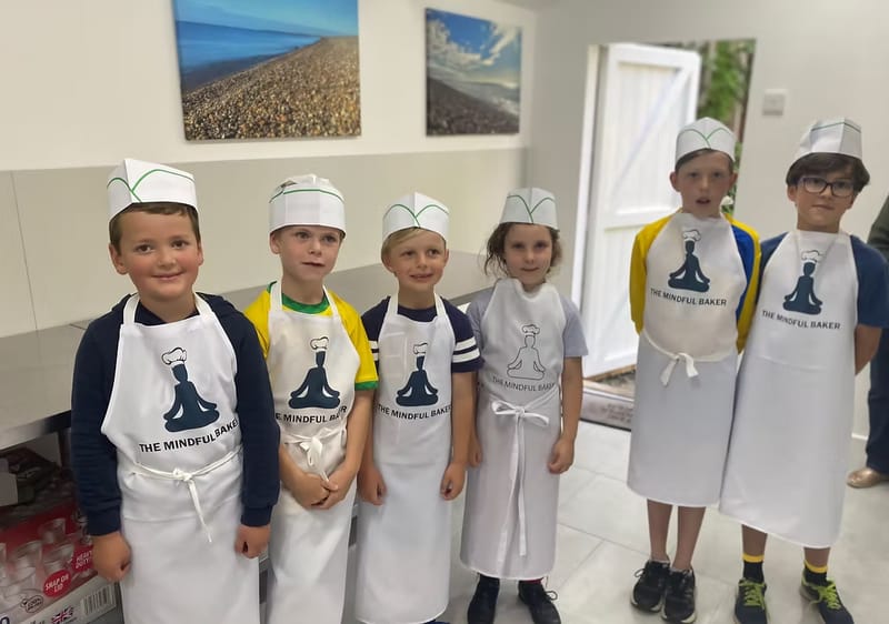Six children lined up in Mindful Baker aprons and paper chef hats, ready to start