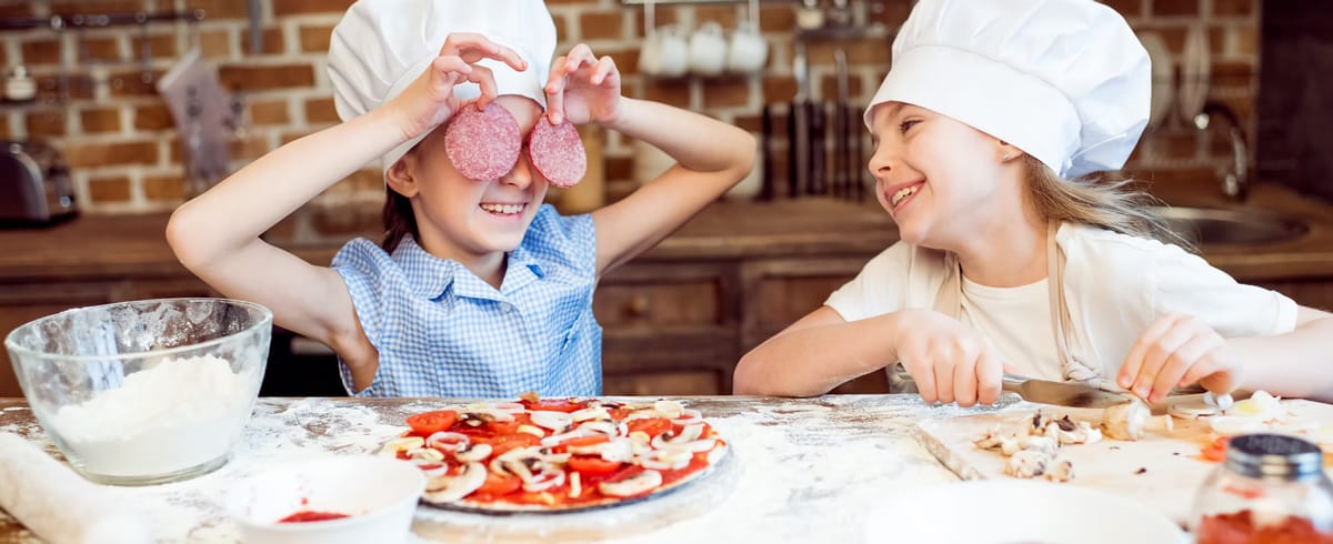 Two girls in chef's hats laughing as one holds salami slices over her eyes while they top a pizza