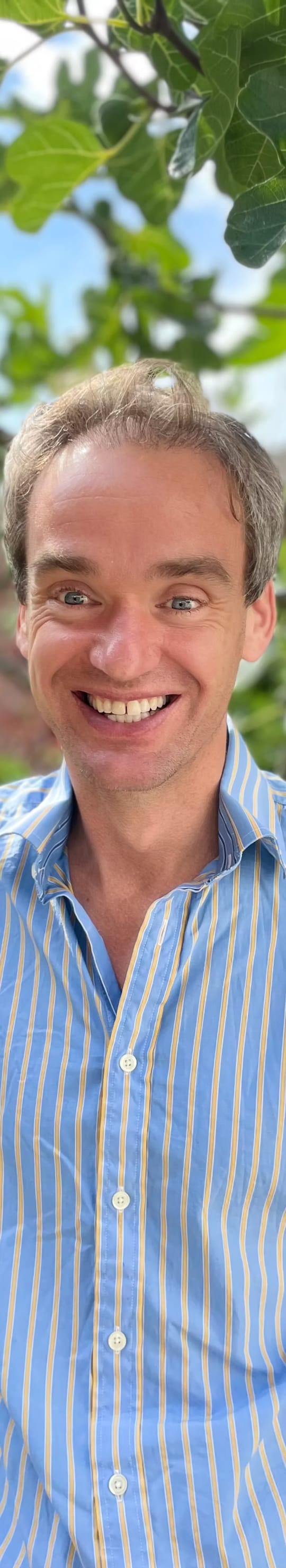 Tim, The Mindful Baker, grinning under a fig tree in a blue and yellow striped shirt