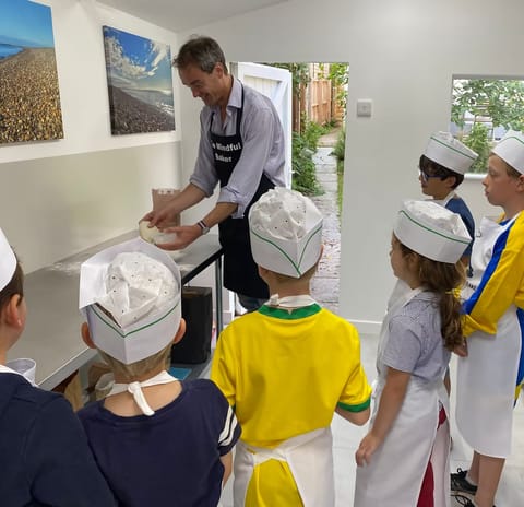Tim, in his Mindful Baker apron, holds up a tub of dough for a group of children in paper chef hats